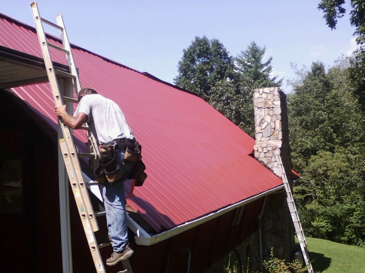 Roofer on a ladder beside a red metal roof and stone chimney — Soffit & Fascia Repair by Roofing Brothers in Princeton Junction